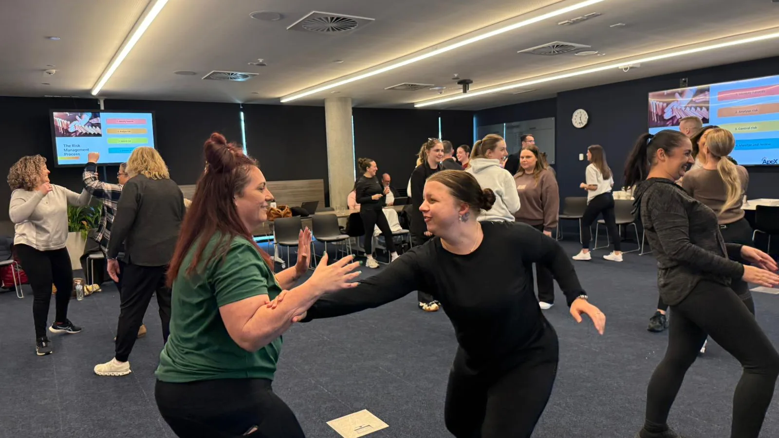 Two participants laugh as they practise breaking free from a grab, with the rest of the team practising in pairs around them during a personal safety workshop